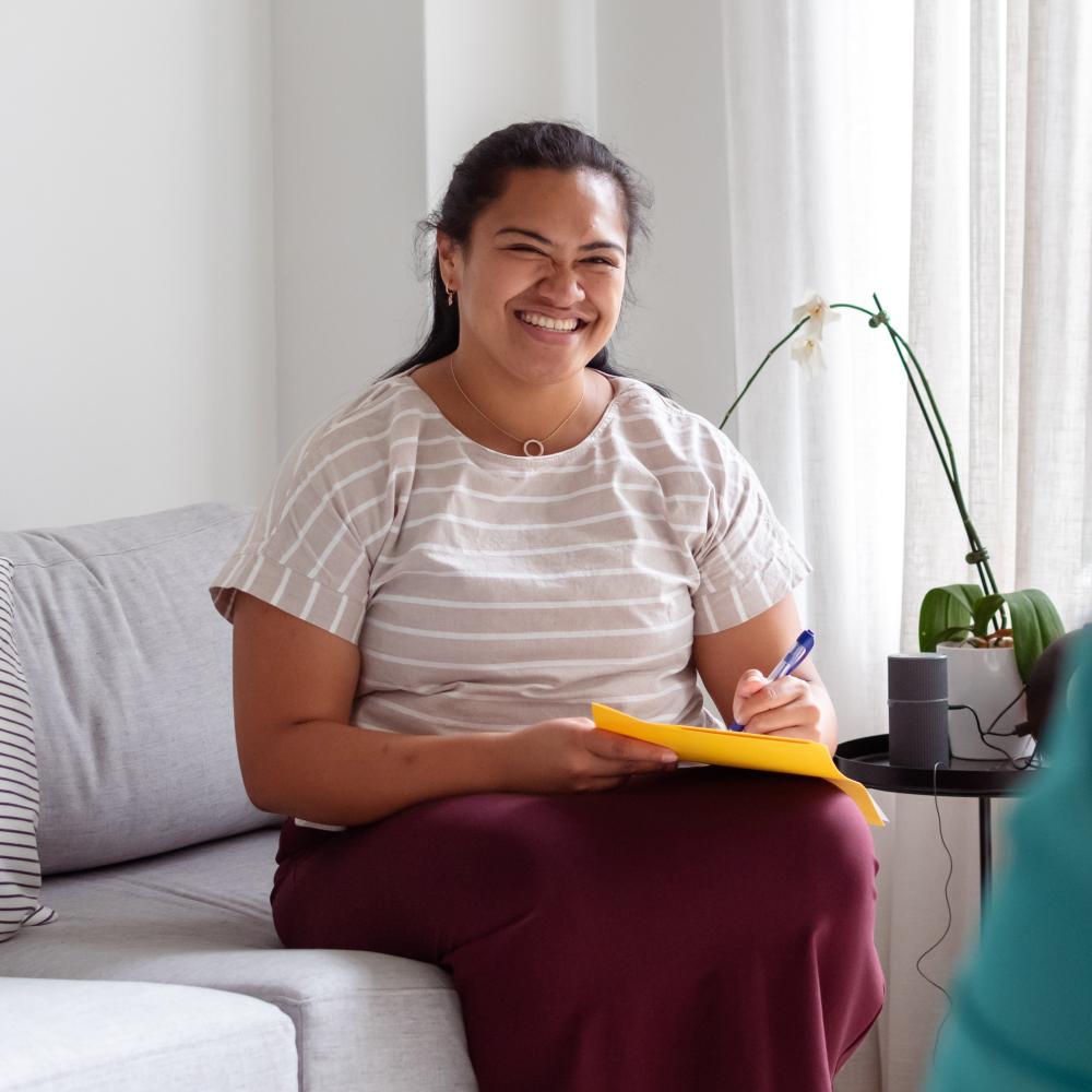 A woman sits and smiles while holding a pad and pen