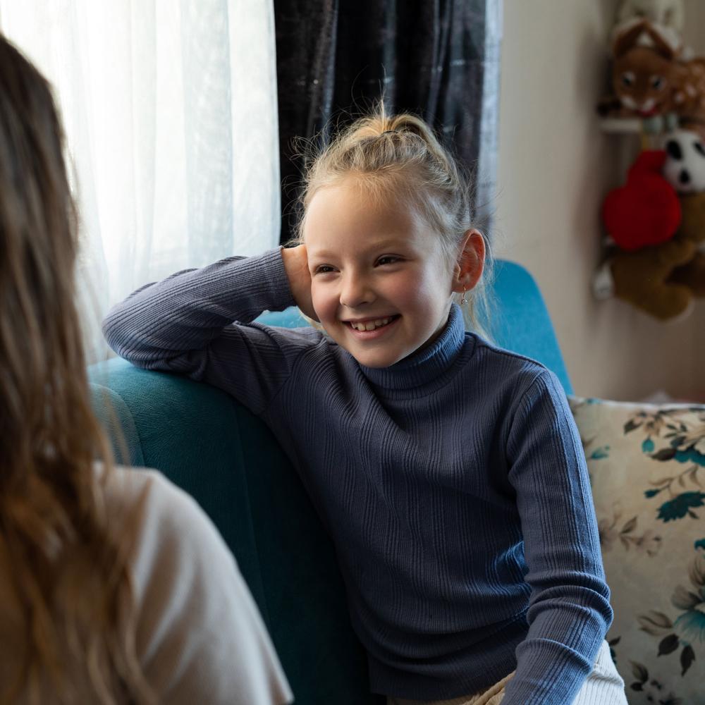 A young girl sits and smiles while talking to a woman